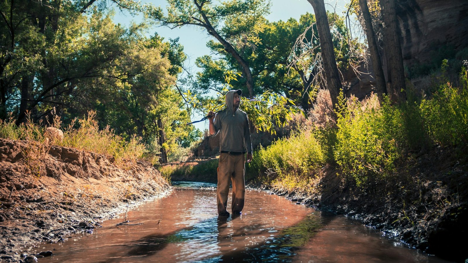 man wearing hooded shirt standing on river surrounded with green trees during daytime